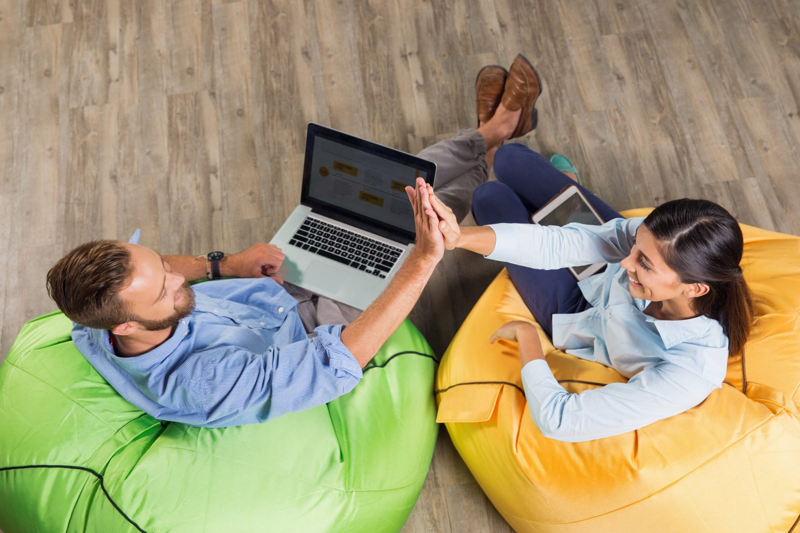 Smiling man and woman sitting on bright beanbag chairs in café, giving high five and working on laptop computer and tablet. Overhead view.